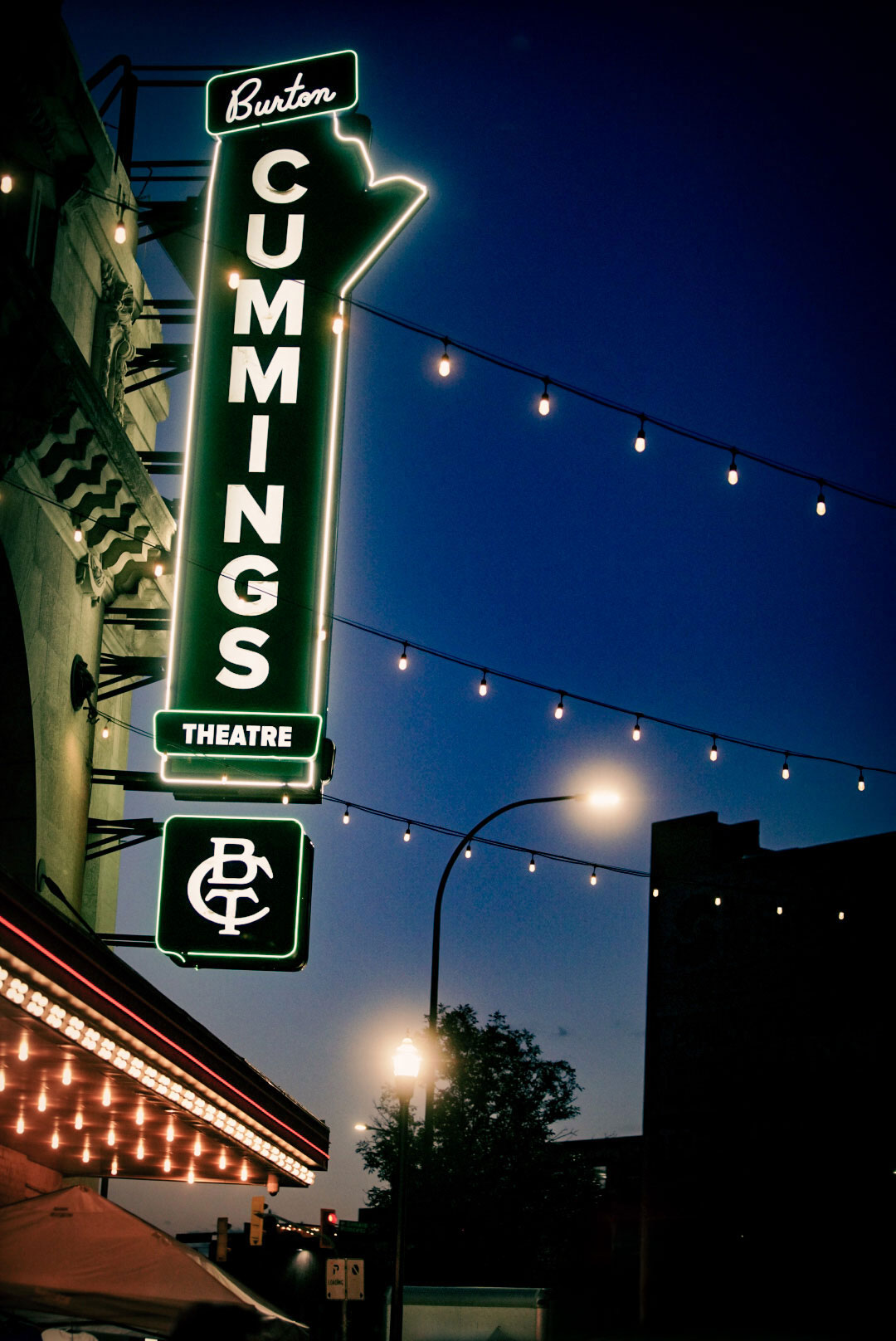 Neon-lit Burton Cummings Theatre sign glowing at dusk, with string lights hanging across the street, creating a vibrant atmosphere in downtown Winnipeg.